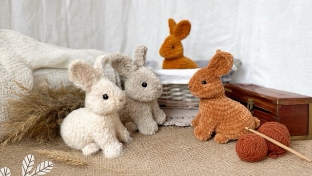 Three handmade crocheted baby rabbits displayed with yarn, a crochet hook, and a basket.